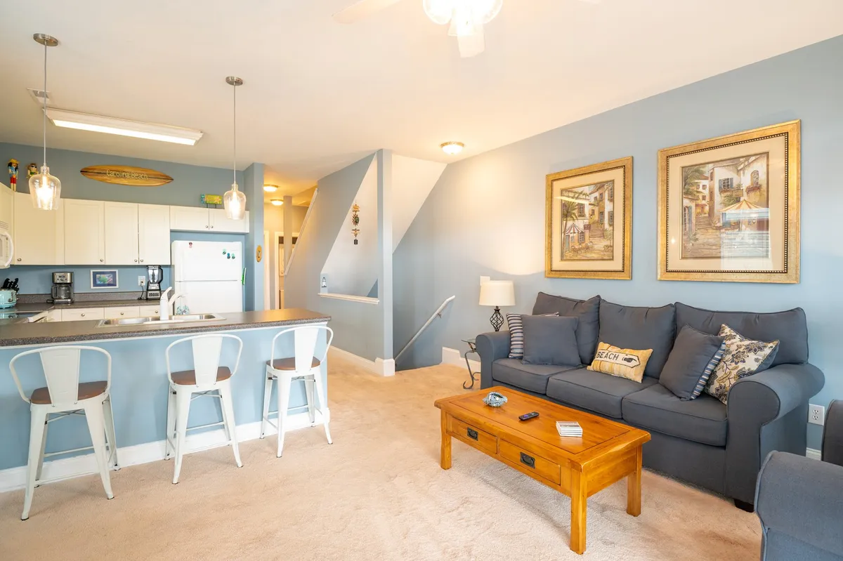 An open-plan living area featuring a comfortable sofa, coffee table, bar stools, and a kitchen area in the background. The space is bright, with artwork on the walls and a welcoming, cheerful vibe. The layout shows the seamless flow between lounging and kitchen spaces, capturing a hub for social gatherings, meals, and movie nights.