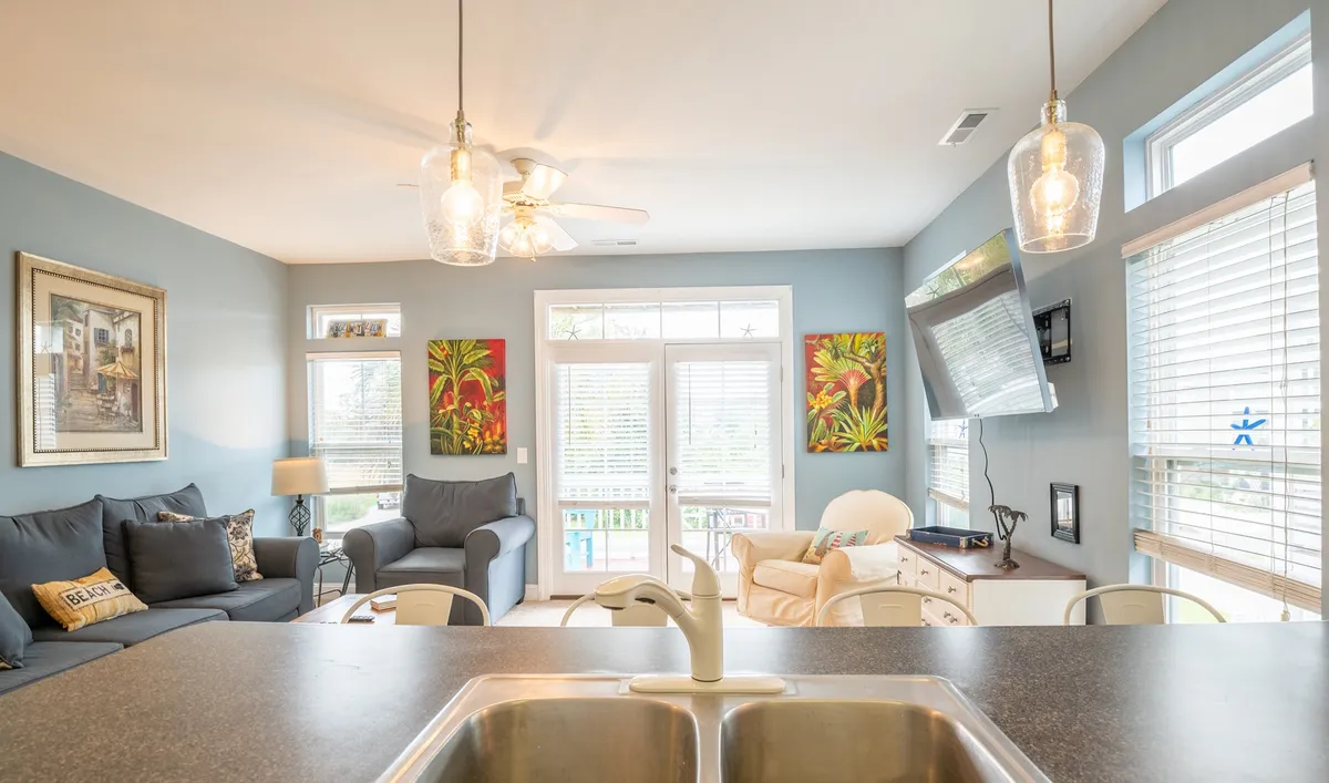 This bright, open living area features a comfortable sofa with accent cushions, stylish artwork, a dining space, and large windows offering views outside. The room is filled with natural light, creating a welcoming environment for socializing, relaxing, or enjoying meals together.
