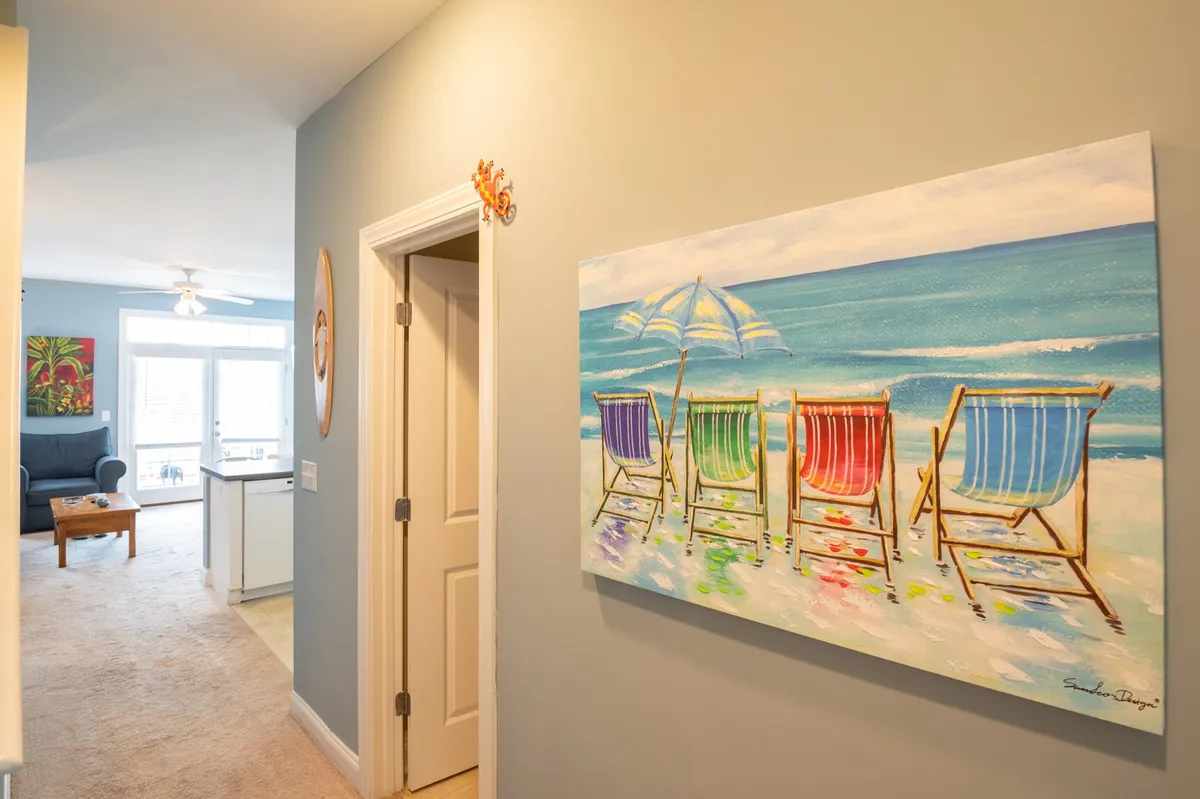 A cheerful transition area with light-colored walls, a vibrant painting of beach chairs, and a view into a connected living space. The open doorway hints at spaciousness and easy flow between rooms, ideal for groups moving freely through the home.