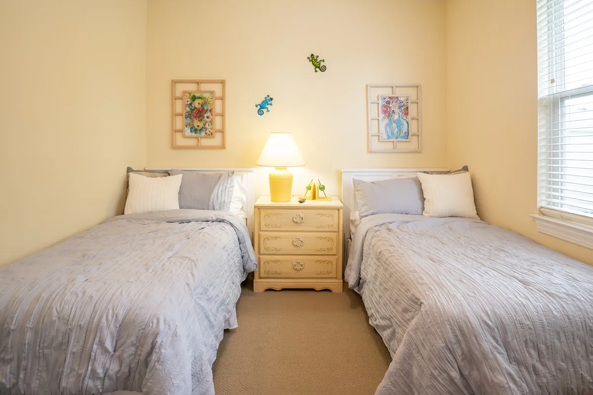 A cheerful bedroom with two twin beds, a shared nightstand with a lamp, and framed wall art. Soft morning light filters in, creating a comfortable environment for both privacy and shared family experiences.