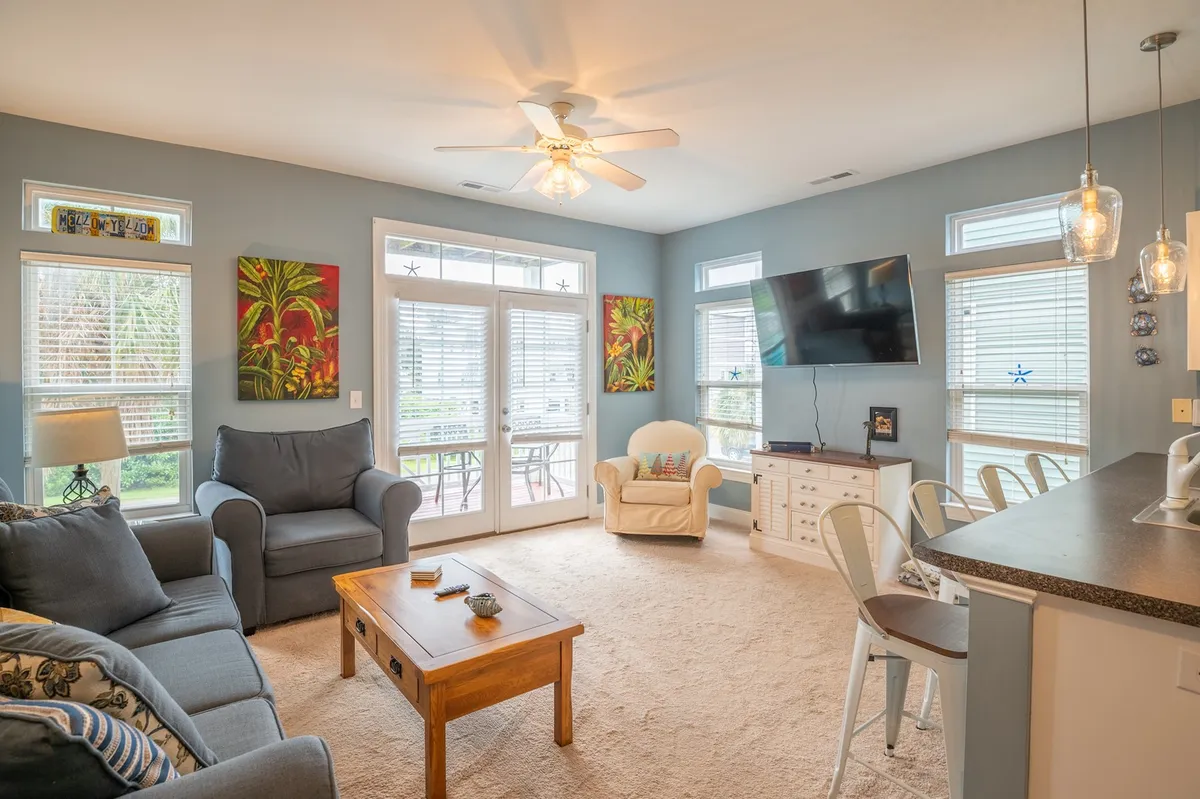 A sunlit living room with a plush sofa, coffee table, colorful artwork on the walls, a television, and large windows that let in plenty of natural light. The space feels inviting and comfortable, ideal for socializing or relaxing.