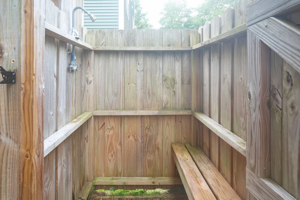 A rustic outdoor shower with wooden walls and a simple bench offers a private, refreshing space to rinse off after a day at the beach. The setup is practical yet blends naturally into the yard’s surroundings.