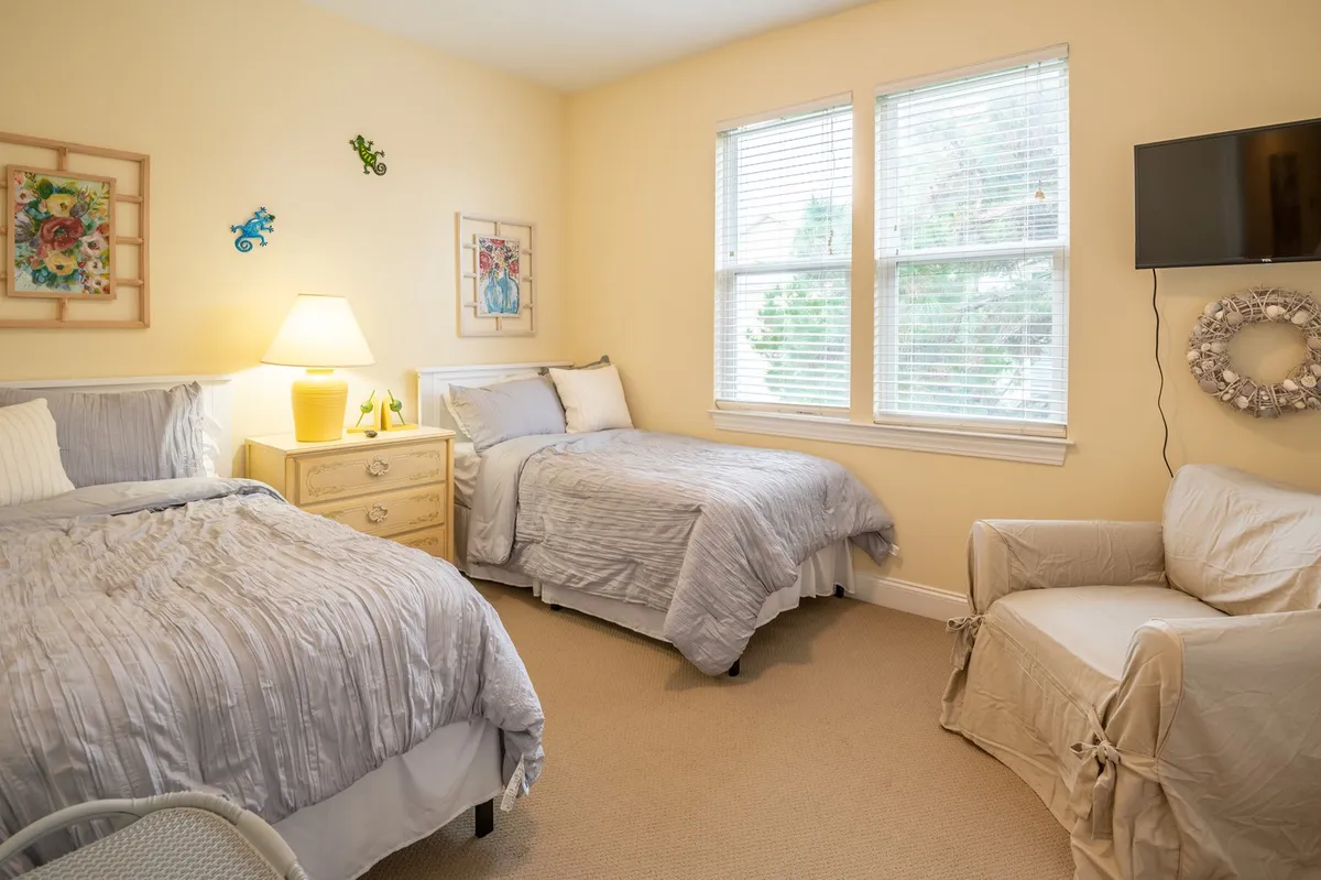 A cozy guest bedroom featuring two beds with gray bedding, a nightstand with a lamp, artwork on the walls, and a comfortable seating area. The room looks inviting and suitable for both family members and friends.