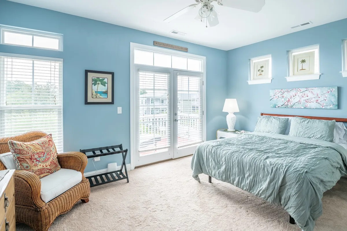 This image shows a bedroom with light blue bedding, a wicker armchair, framed artwork, and double doors leading to a private balcony or sun deck. The room is filled with natural light, creating a tranquil and serene atmosphere.