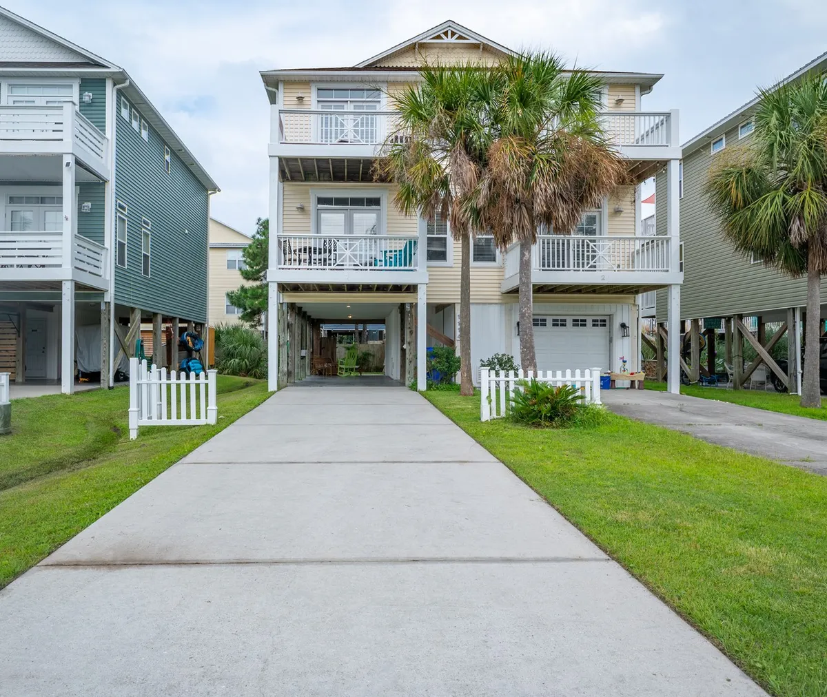 The image showcases a charming two-story yellow beach house framed by swaying palm trees, a classic white picket fence, and a spacious driveway. The inviting front porch and upper balcony hint at relaxing mornings with coffee or serene sunset evenings, while the tranquil setting exudes laid-back coastal charm.