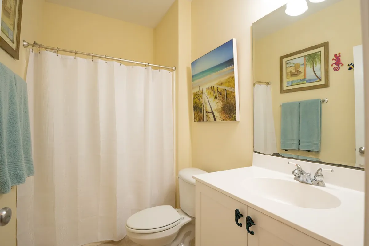 A clean, modern bathroom with a white shower curtain, vanity and sink, towels, and beach-themed artwork. The space feels relaxing and refreshing, ideal for self-care routines.