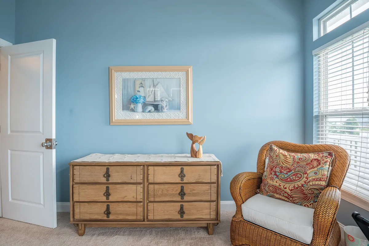 A cozy, sunlit nook featuring a wooden dresser, framed artwork, and a rattan chair with a plush cushion. The space feels welcoming and is ideal for enjoying a morning coffee or having relaxed conversations with friends.