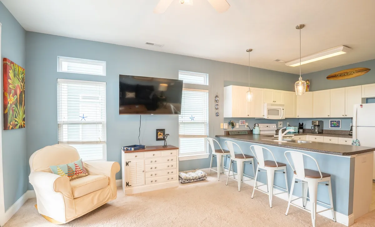 A cozy interior featuring a white armchair, a kitchen island with bar stools, a large flat-screen TV, and vibrant wall art. The space is designed for relaxation, conversation, and casual meals or entertainment.